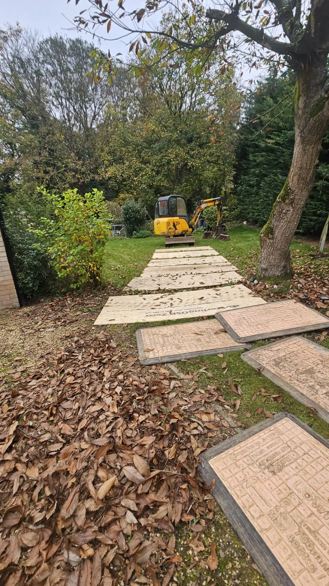 Mini excavator on grass with temporary ground protection and drainage access covers, showing active groundworks.