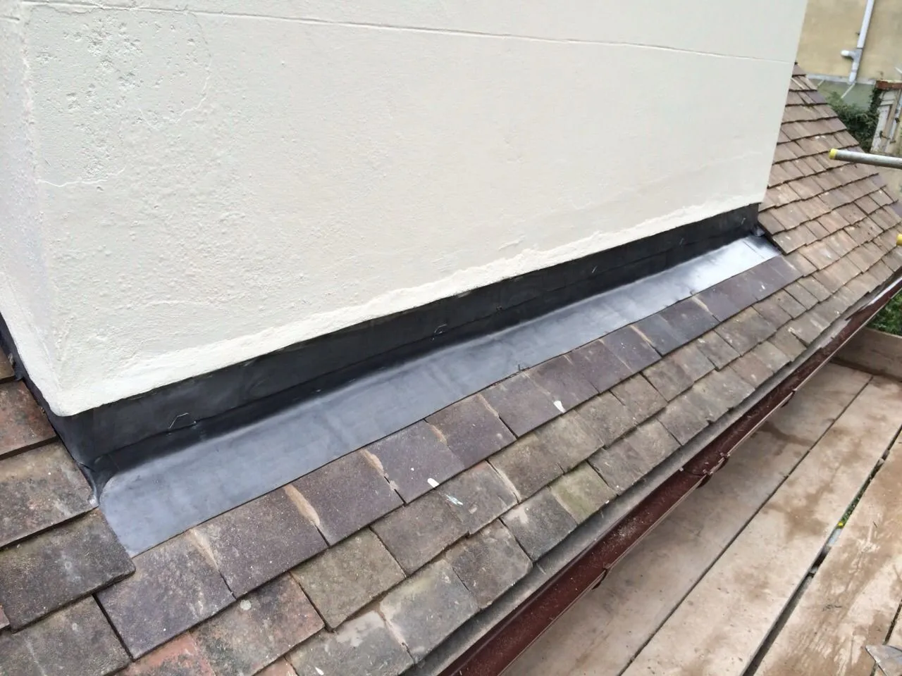 Close-up of newly installed lead flashing on a tiled roof next to a painted wall or chimney.