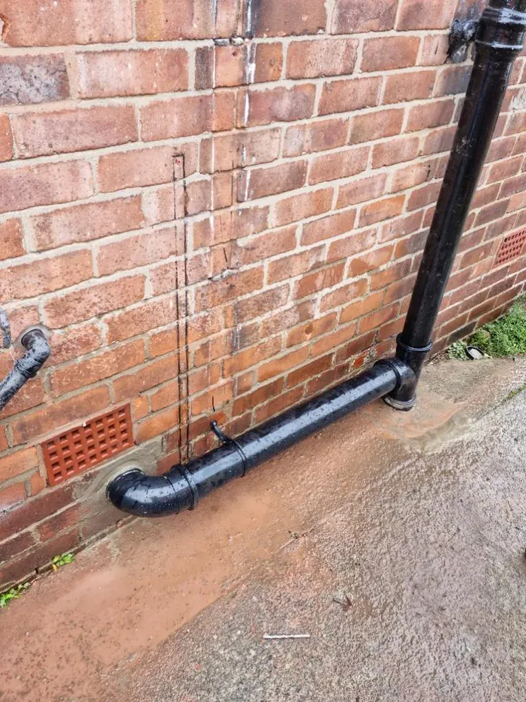 Blocked drain pipe with water pooling outside a brick building wall.