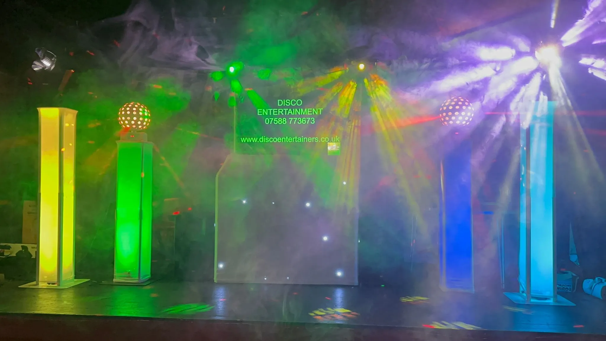 Dynamic DJ lighting display with green, blue, and purple lights, haze, and illuminated pillars at a lively event.