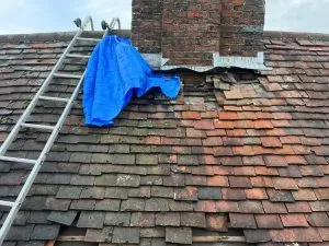 Emergency roof repair with missing tiles, a temporary blue tarp over the damaged section, and a ladder next to a chimney.