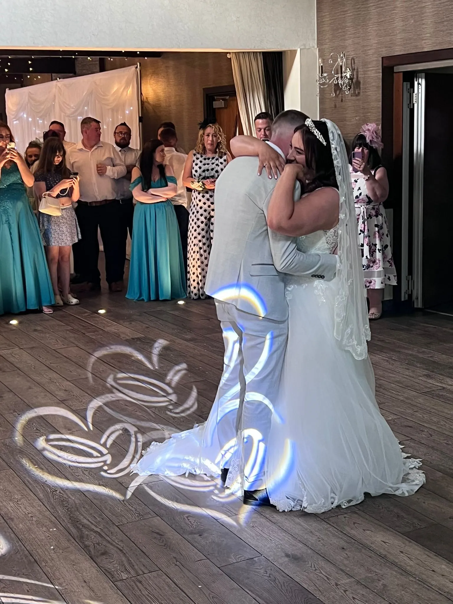 A bride in a white gown and a groom in a light suit sharing their magical first dance, surrounded by admiring guests. Projected hearts and rings illuminate the dancefloor beneath them, creating an intimate and joyful atmosphere.