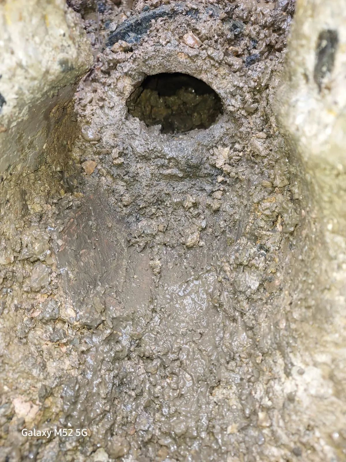 Close-up of a dark, exposed drain pipe opening in muddy, excavated ground with small stones.