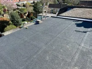 An aerial view of a clean, dark grey flat roof, showing its smooth surface and the surrounding residential area with gardens and other rooftops under a clear sky.