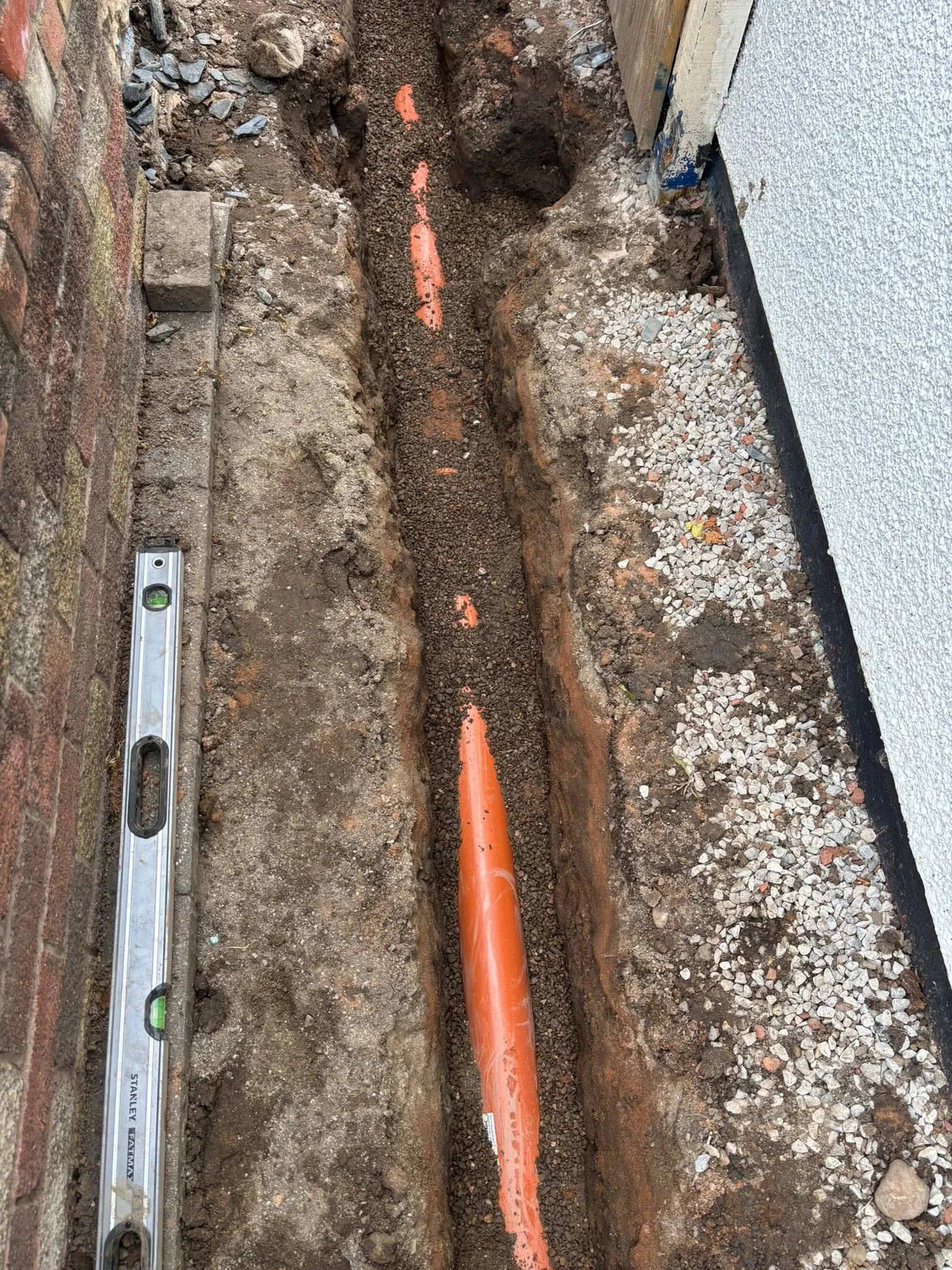 Trench with orange drainage pipes being installed next to a brick wall and rendered house wall