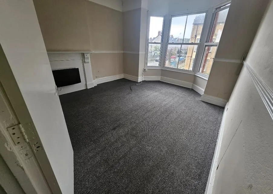 A picture of a cleared, unfurnished residential room with a large bay window, a decorative fireplace, and clean dark grey carpet.