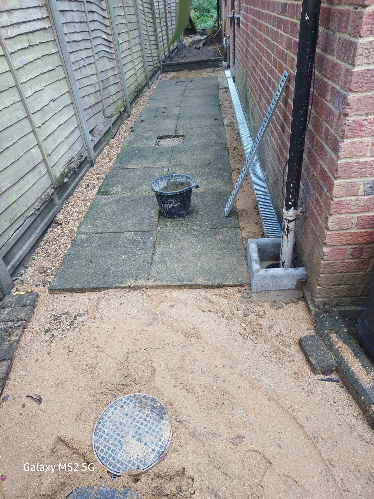 Drainage installation showing a new channel drain, downpipe gulley, and a manhole cover on sandy ground next to a building.
