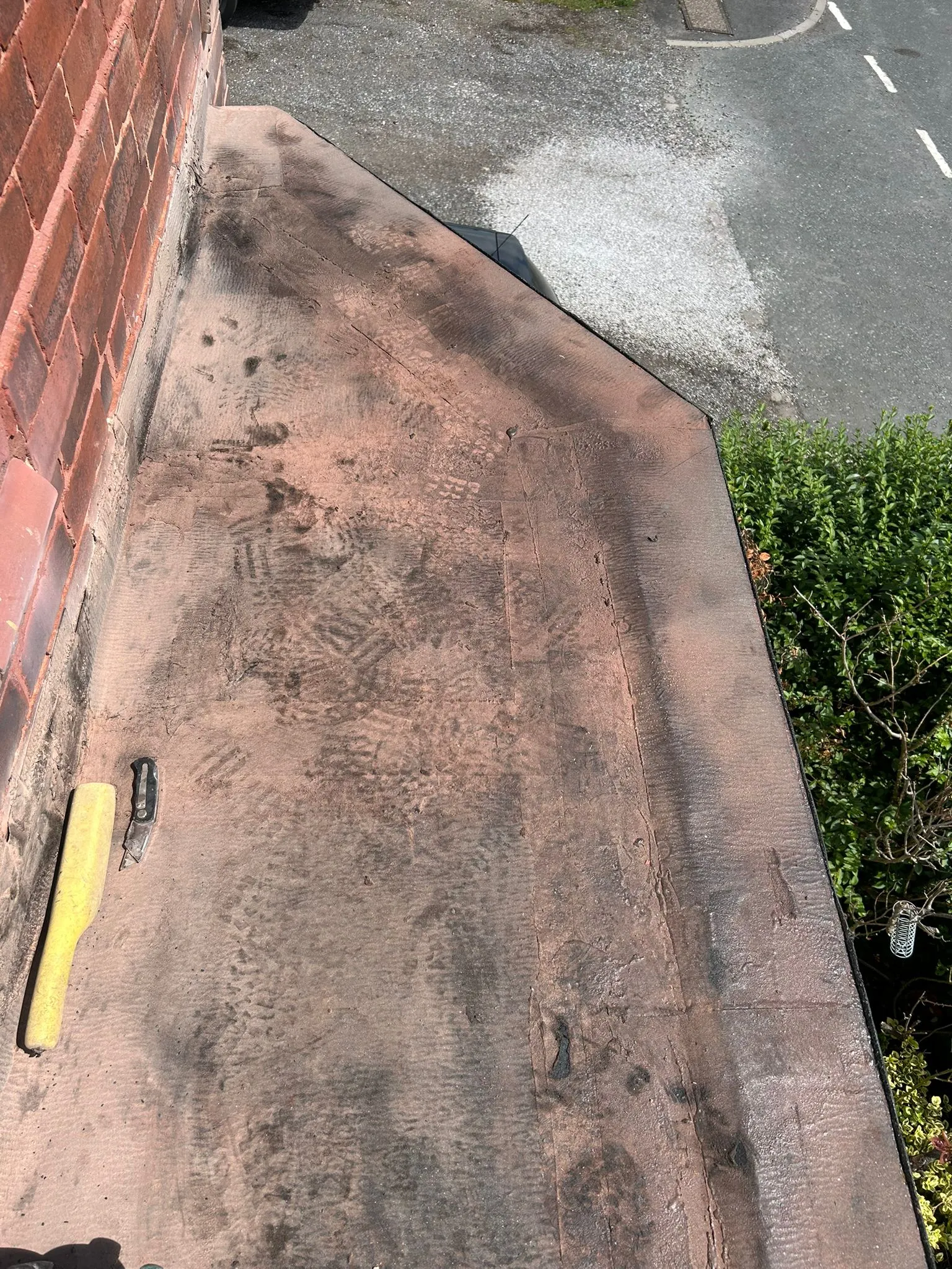 A brown torch-on felt flat roof with a utility knife and yellow tool, indicating ongoing or pre-repair work.