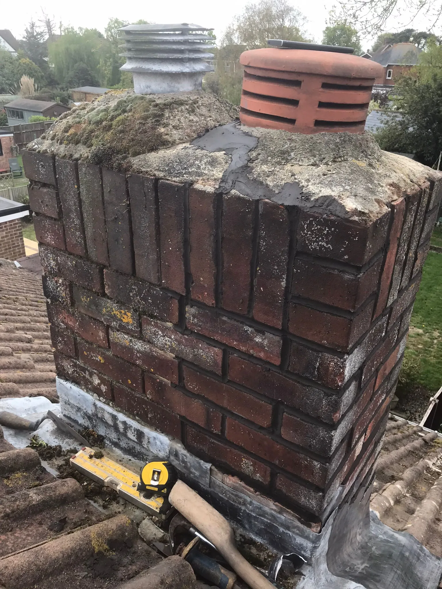 A weathered brick chimney stack on a tiled roof with moss, worn mortar, lead flashing, and two distinct chimney pots, indicating a need for repair or maintenance.