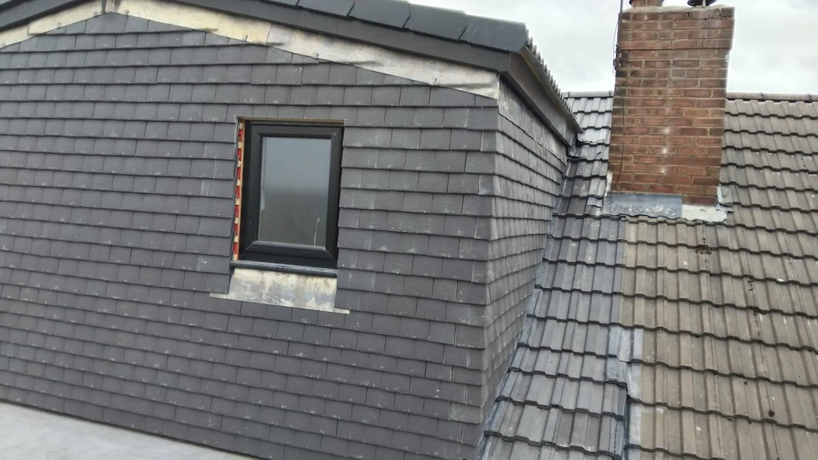 A picture showing a new dark grey tiled dormer on a roof next to an older tiled section and a brick chimney.