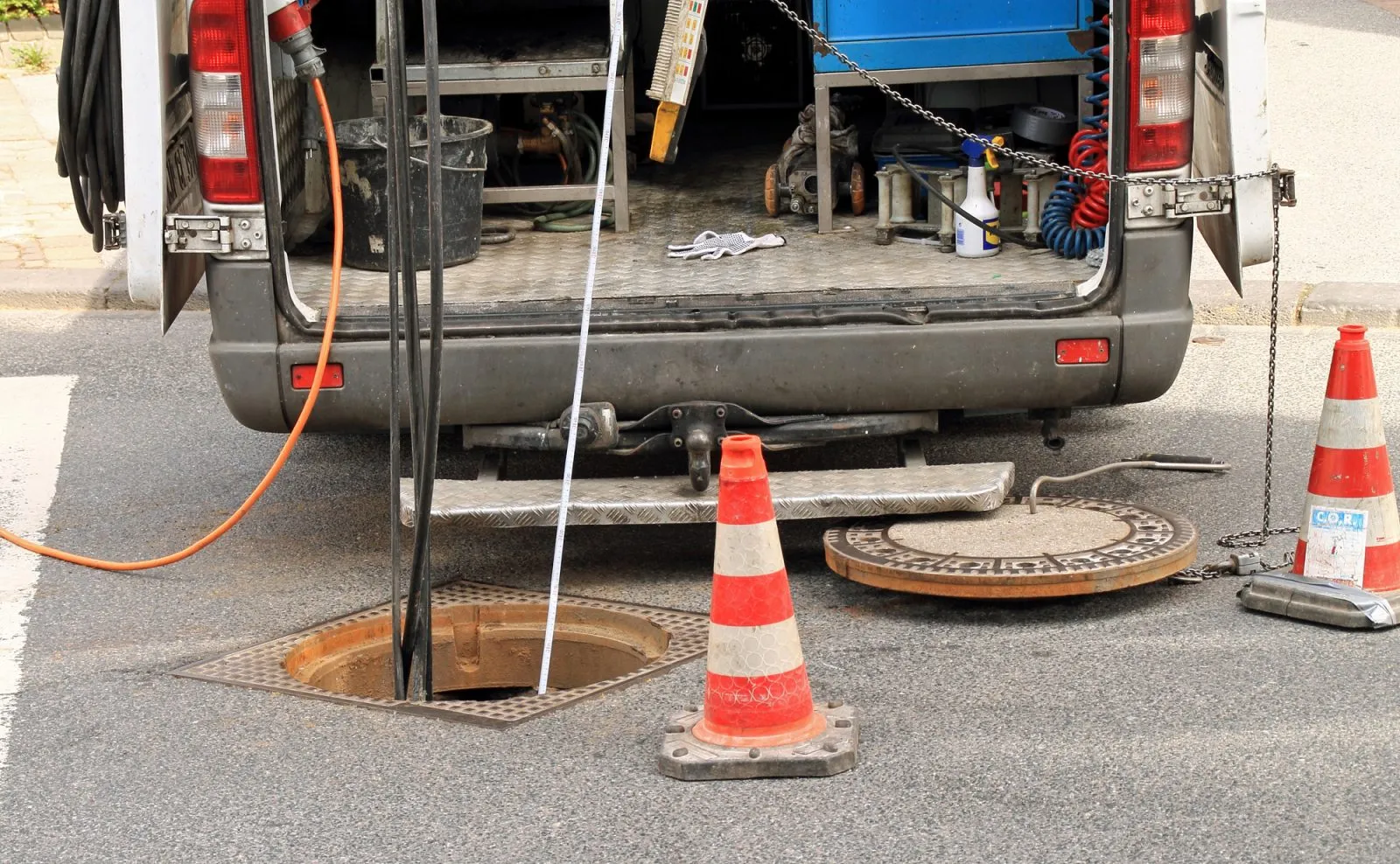 Drainage engineers' van and equipment at an open manhole on a road, performing outdoor drain unblocking or repair.