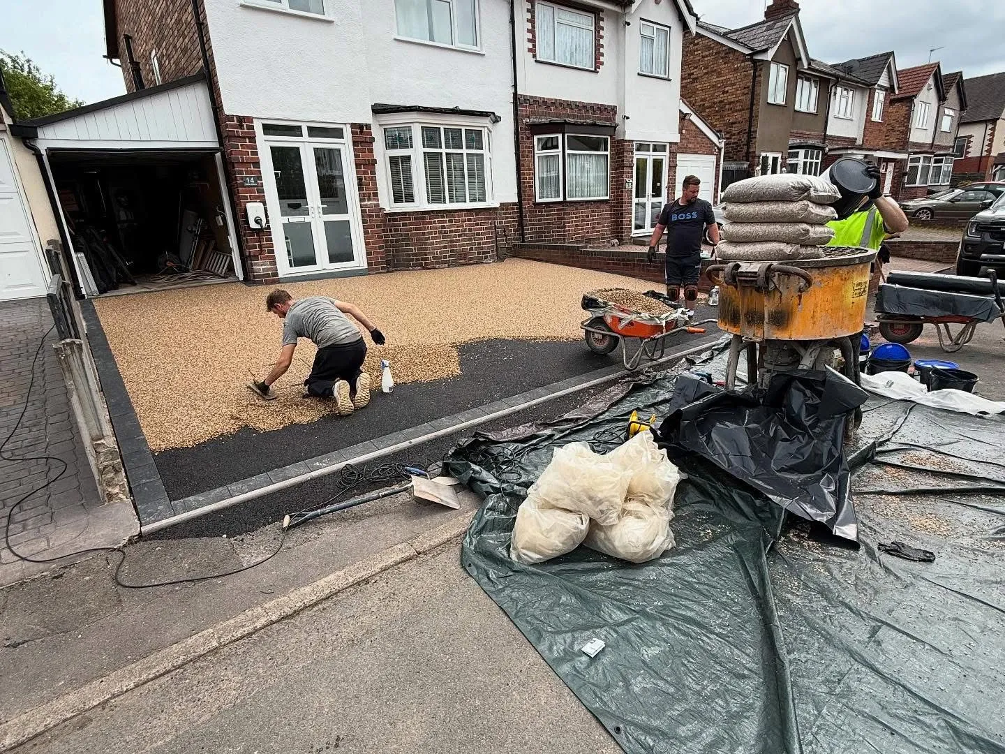 Workers trowelling a new permeable resin-bound driveway with golden aggregates, a mixer, and materials on site for Top Pave Driveways LTD.