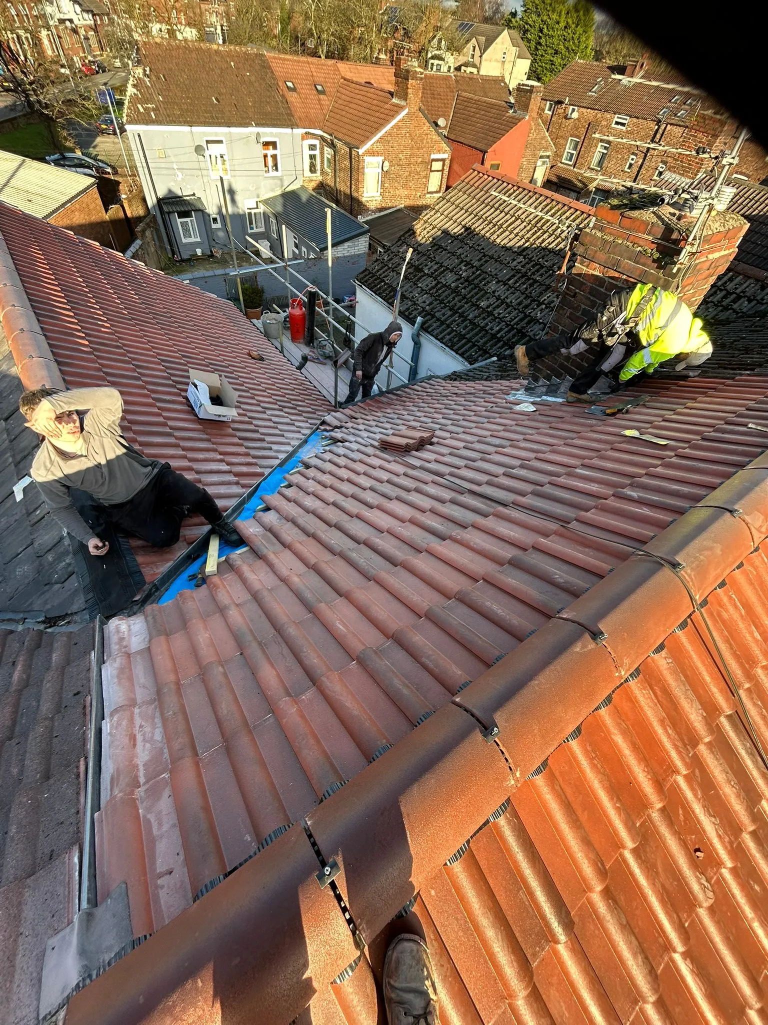 Roofers from M. Woodward Roofing installing or replacing red clay tiles on a pitched roof, with one roofer working near a chimney and another resting, showcasing their expertise in roof repairs and installations.