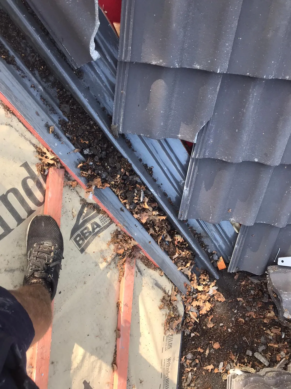 Roof gully or valley filled with autumn leaves, dirt, and debris next to dark grey roof tiles and roofing underlay, showing a blockage.