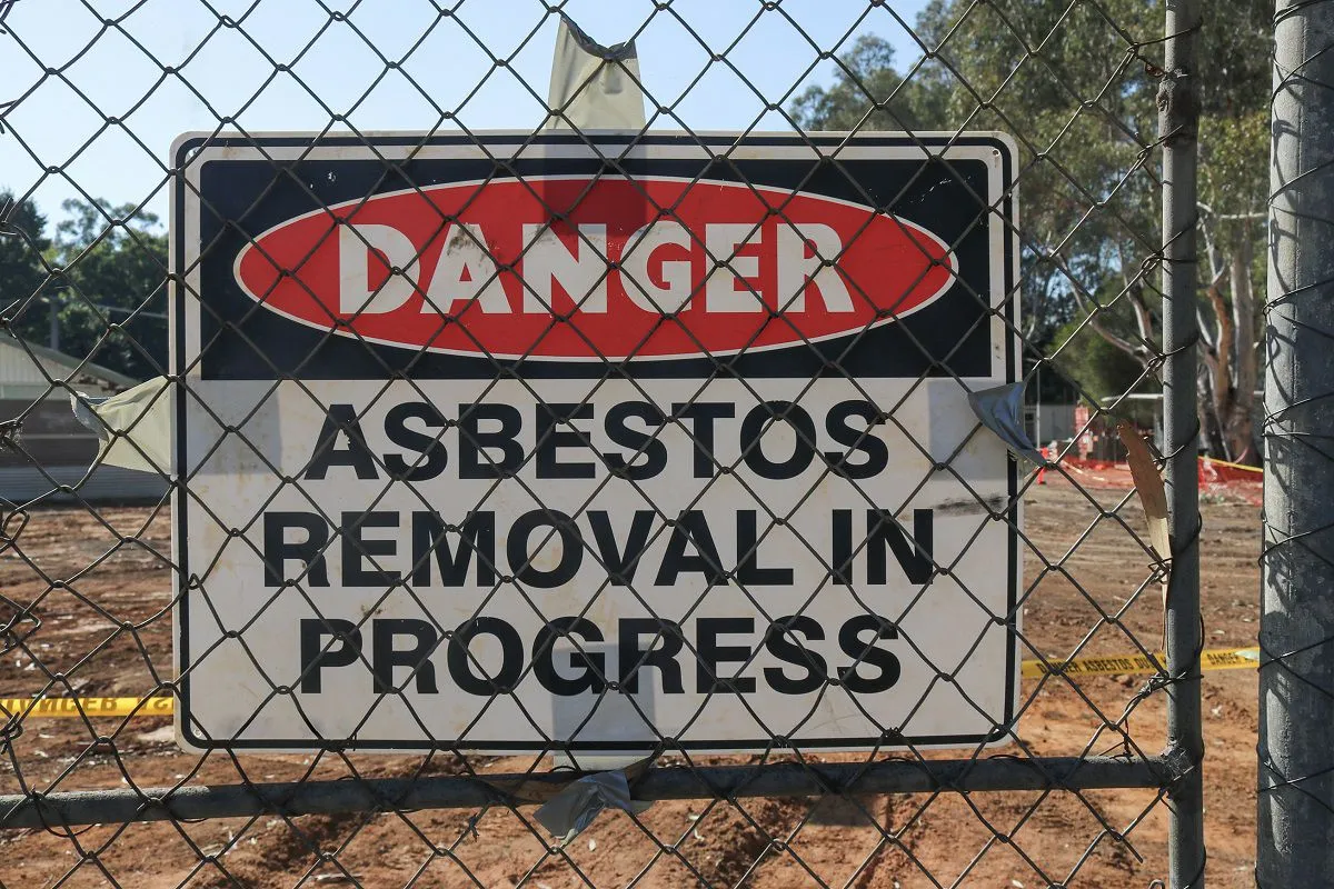 A danger sign on a chain-link fence that reads 'DANGER ASBESTOS REMOVAL IN PROGRESS'.