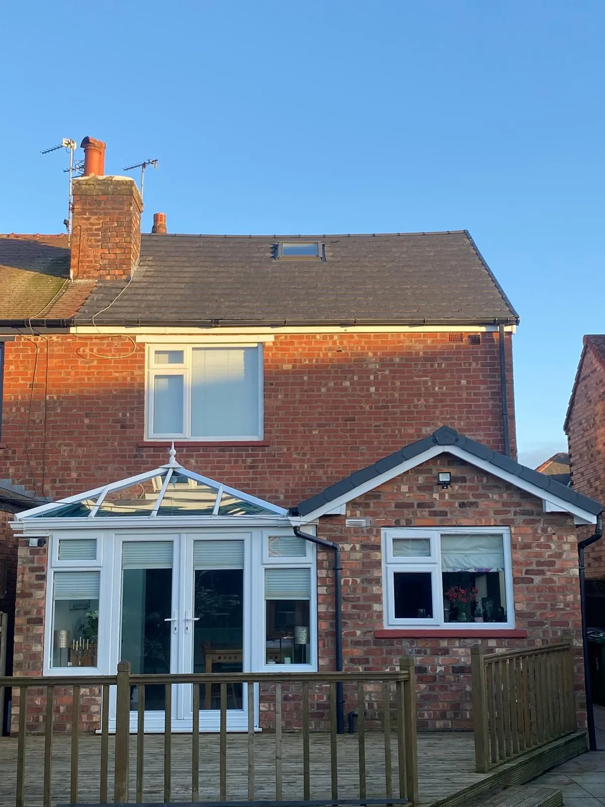 Rear view of a brick house with a new dark grey tiled pitched roof, a Velux window, a white conservatory, and a tiled extension.