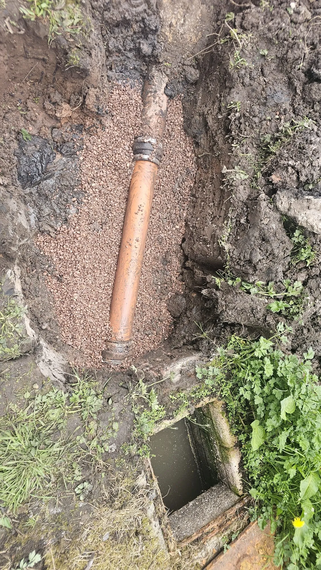 An orange drainage pipe in an excavated trench with gravel, beside an open drain gully.