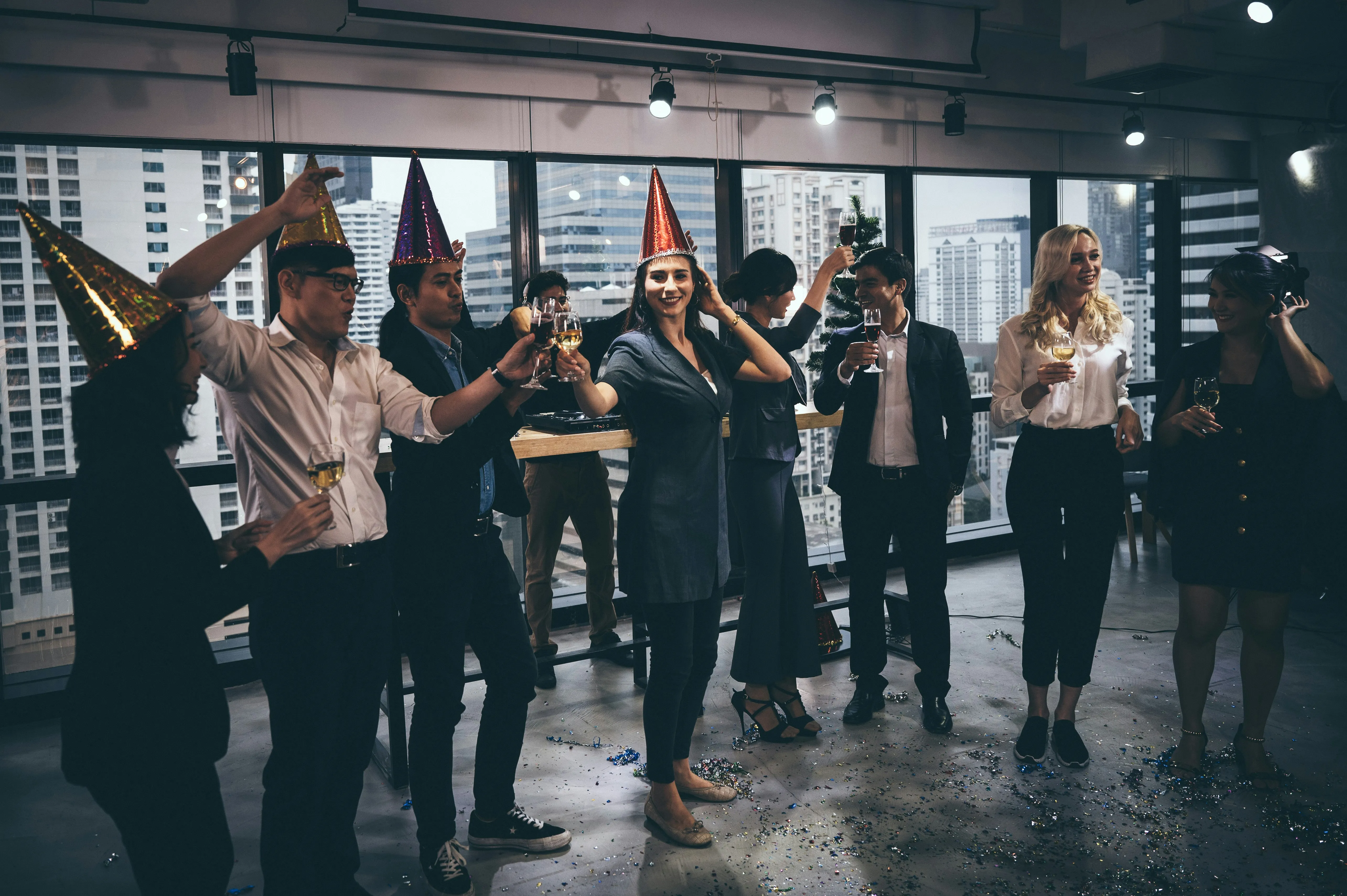 A group of colleagues or friends celebrating at an indoor party with party hats, drinks, and confetti, with a DJ deck and city view in the background.