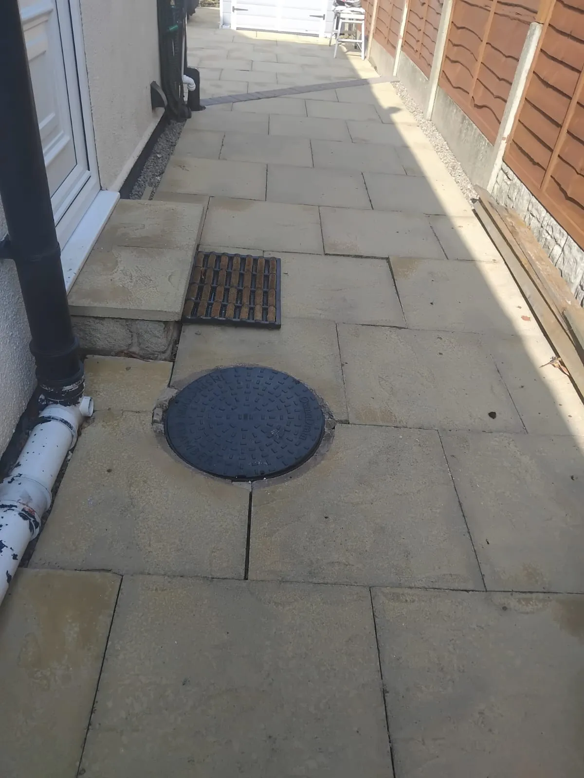 Residential drainage system with a manhole cover, gully, and downpipe on a paved pathway next to a house.