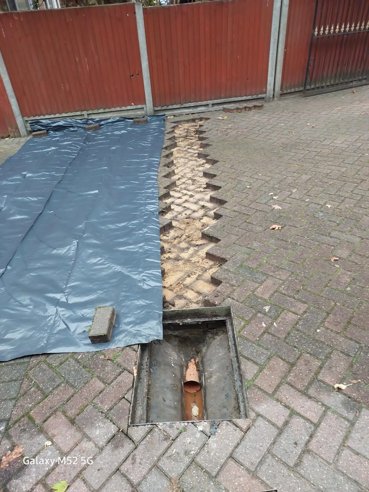 A residential block-paved driveway with a section of paving removed for drainage work, revealing an open manhole with a pipe and a dug trench, partly covered by a blue tarp.