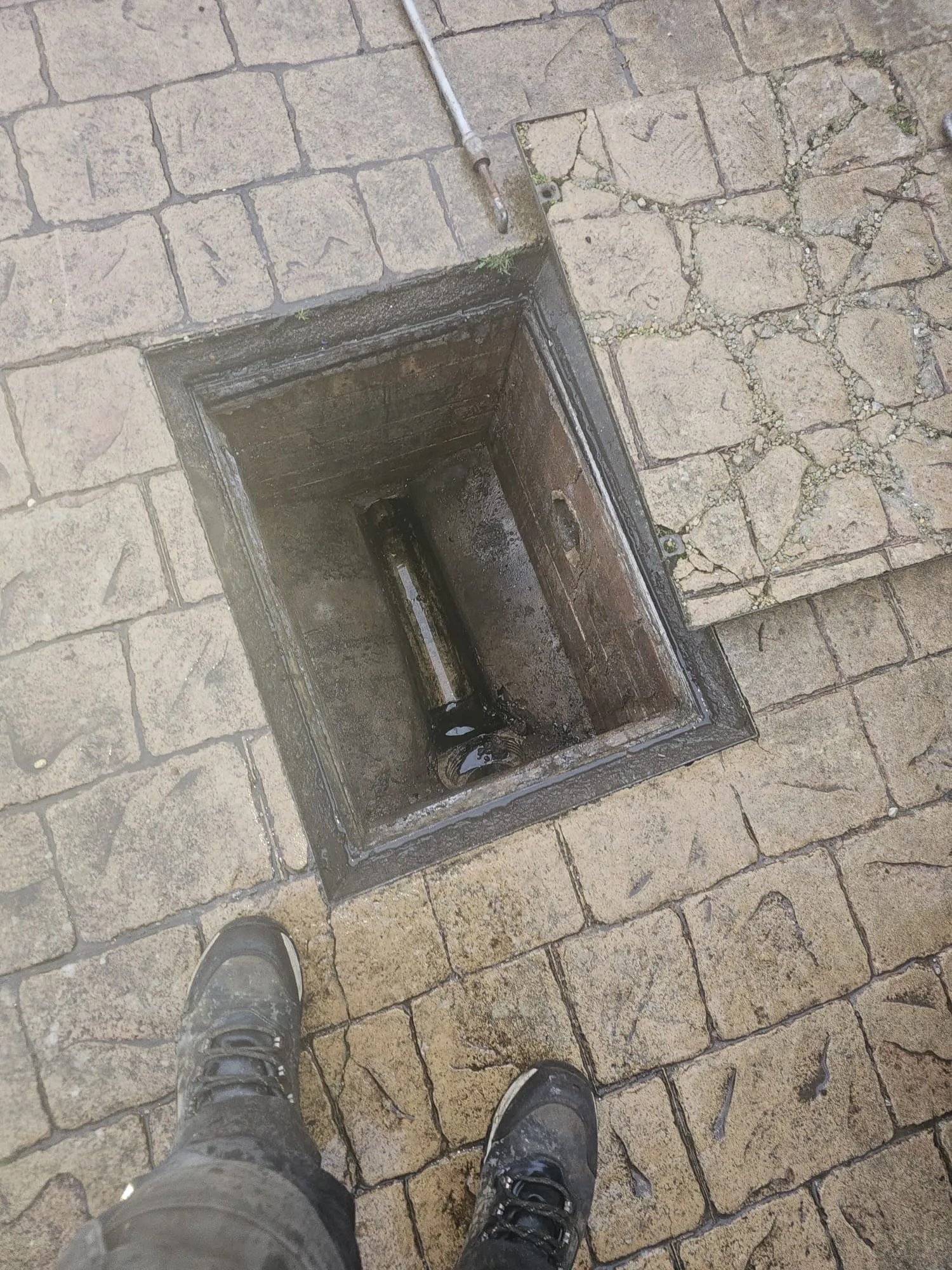 Open manhole access to a potentially blocked drain with standing water on a paved patio