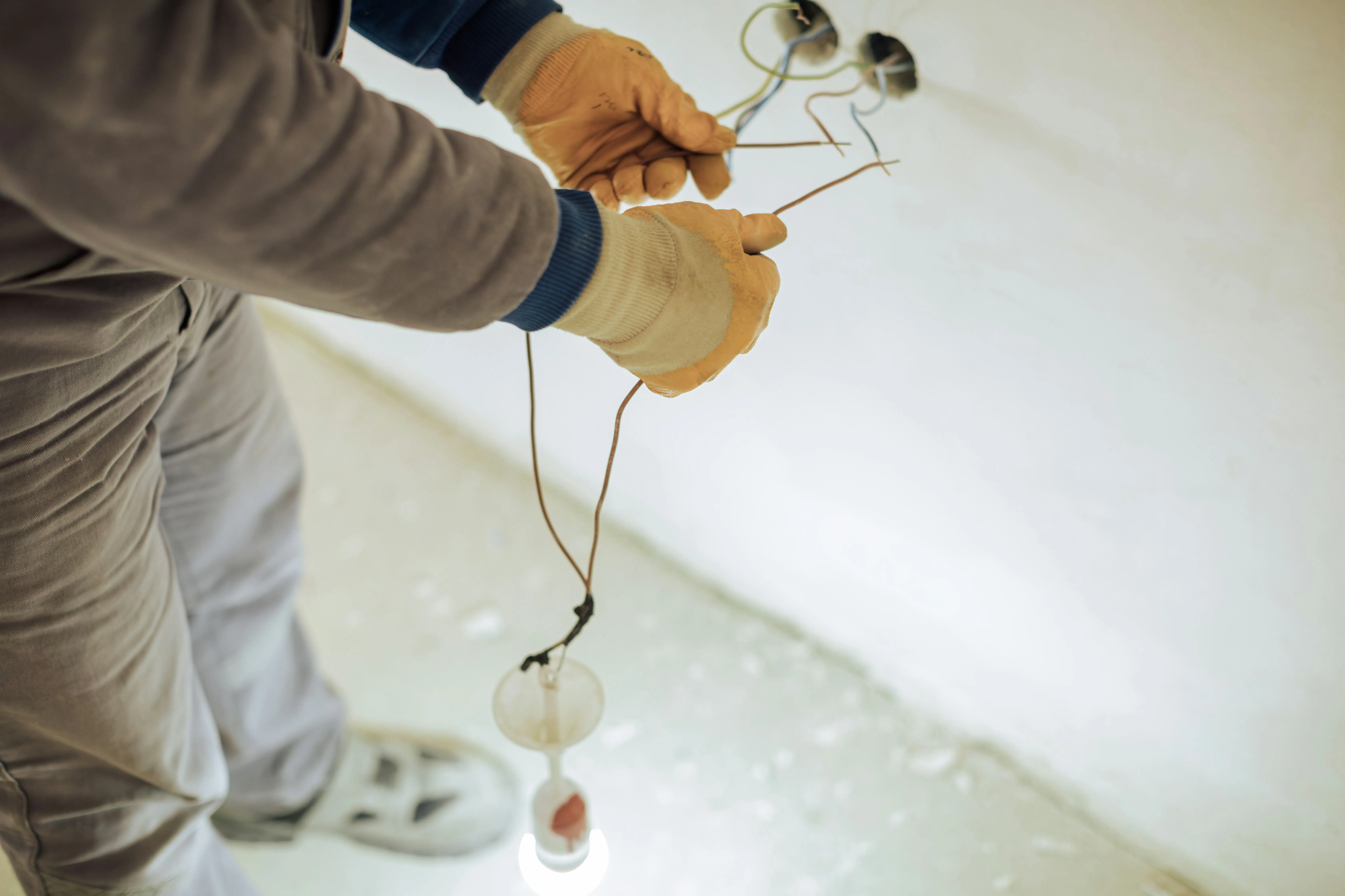 Close-up of an electrician's hands in yellow gloves, installing new electrical wiring for a light fixture in a wall or ceiling during construction, with a temporary bulb illuminating the work area.
