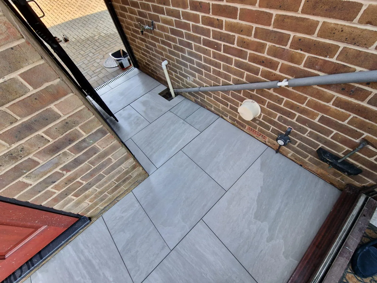 Newly laid grey patio slabs creating a pathway alongside a brick wall, with a sleek linear drainage system connecting to an outdoor block-paved area.