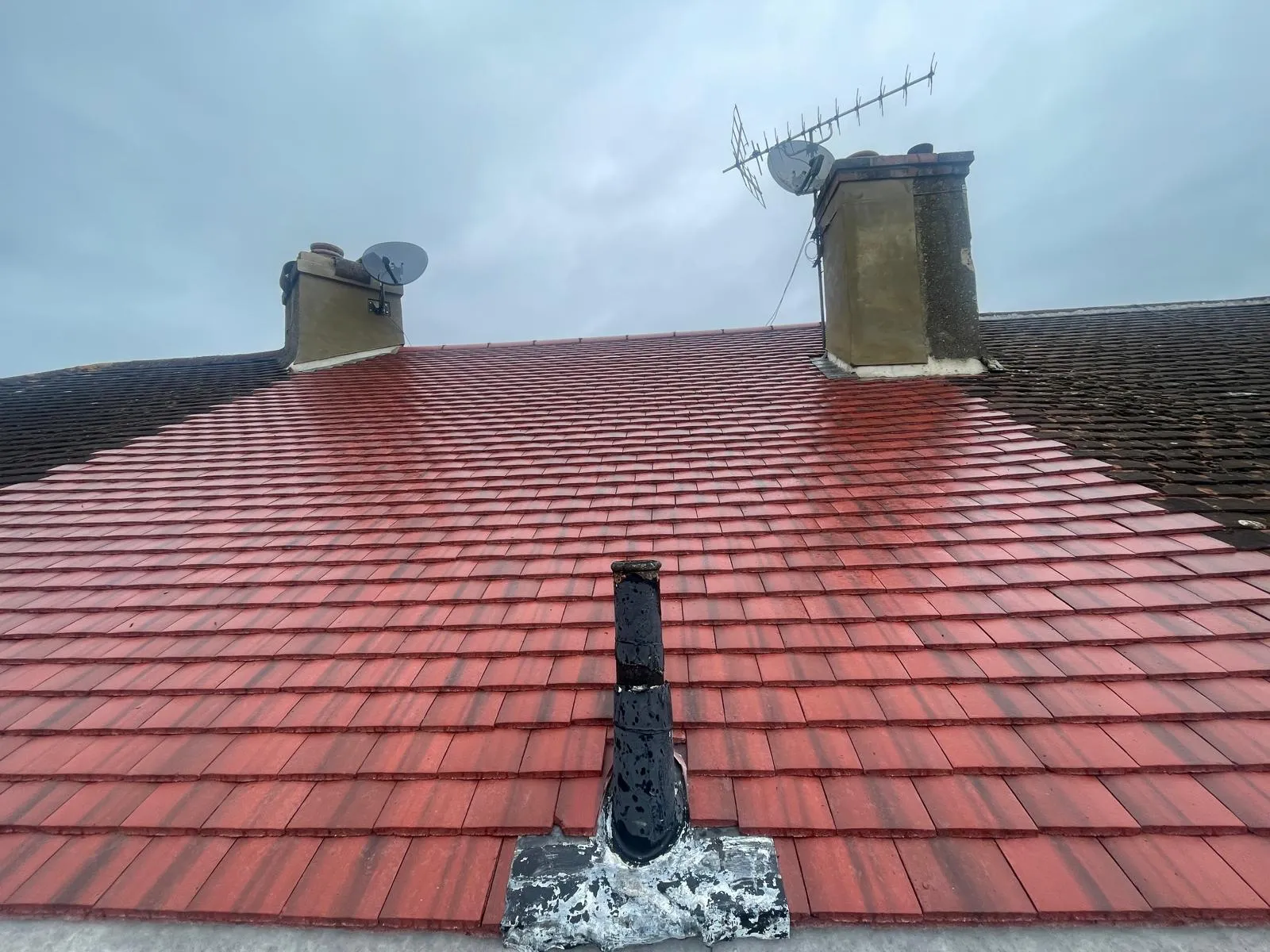 A newly installed or cleaned section of red tiled roof next to older tiles, with two chimneys, satellite dishes, and a TV aerial visible.