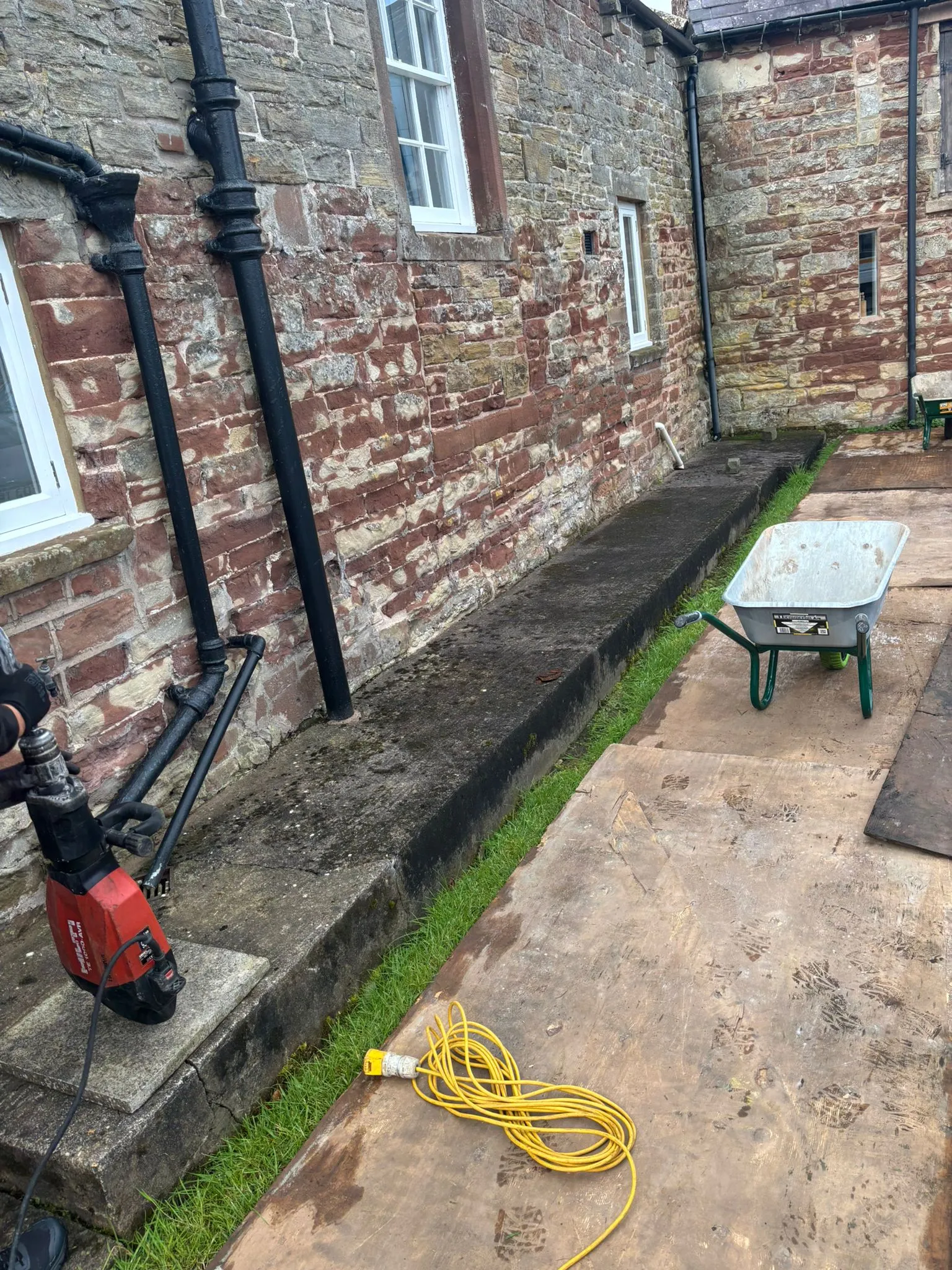 A jackhammer and wheelbarrow on a construction site next to a stone building with black drainpipes, suggesting groundwork or drain excavation.