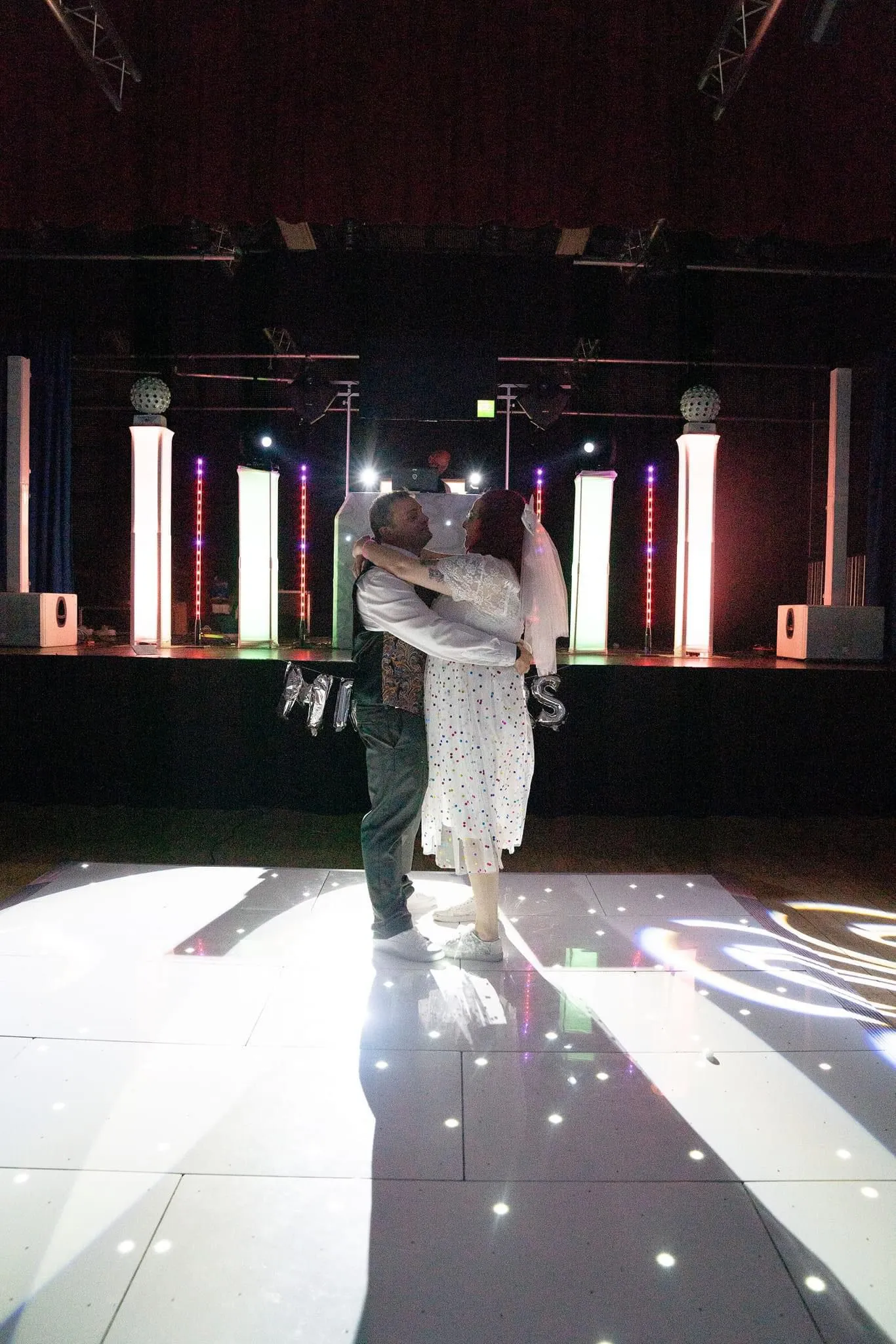 A wedding couple slow dancing on a white LED dancefloor with dynamic lighting from a professional DJ setup in the background, showcasing Disco Entertainers' services.