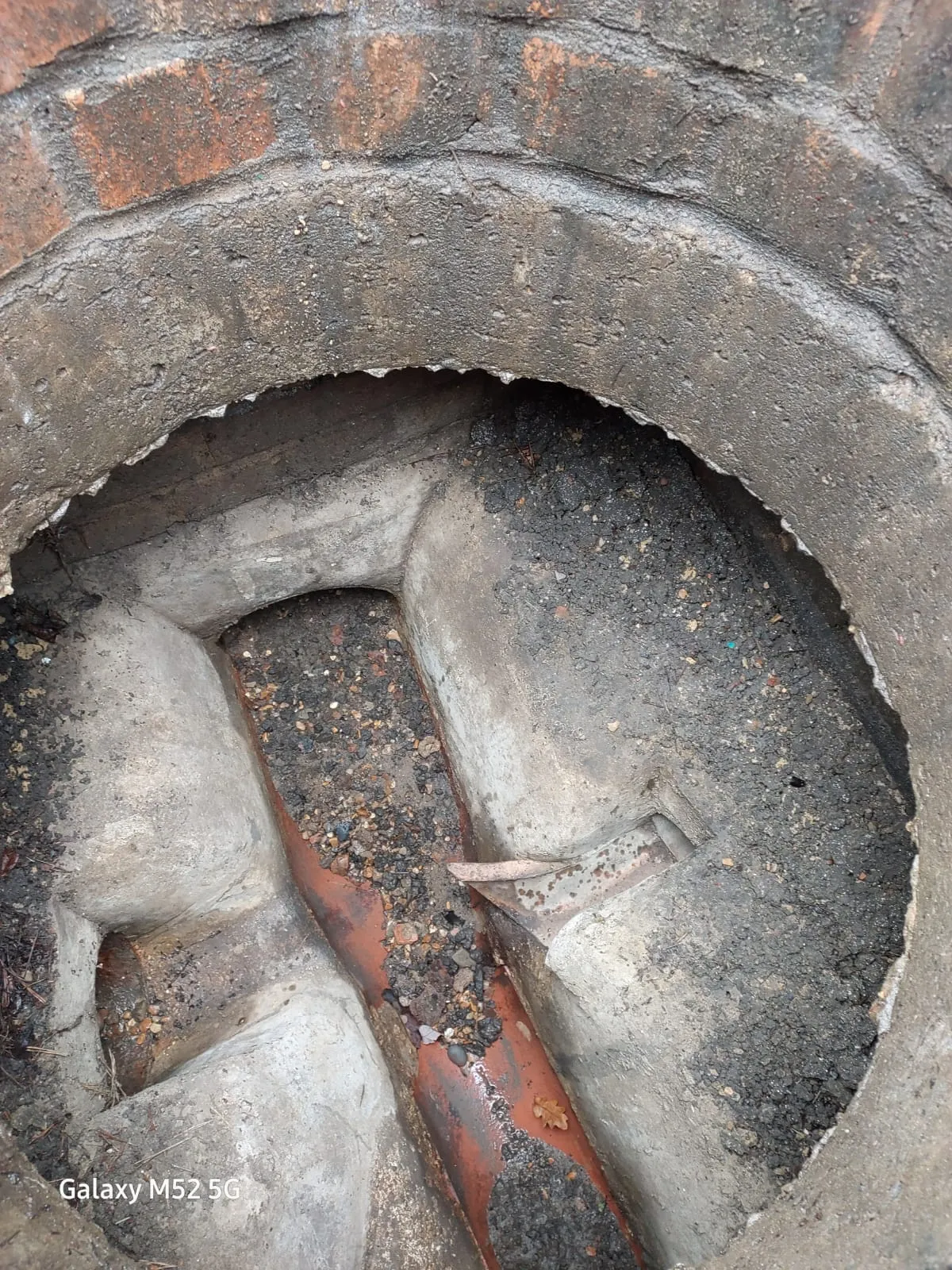 A close-up view inside a brick-lined drainage manhole with a concrete channel filled with dirt, gravel, and water, suggesting a blockage.