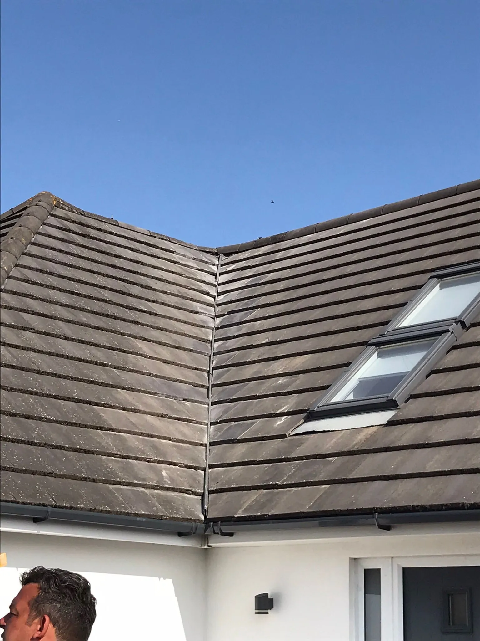 Modern pitched tiled roof with a roof valley, Velux window, and dark grey guttering under a clear blue sky.