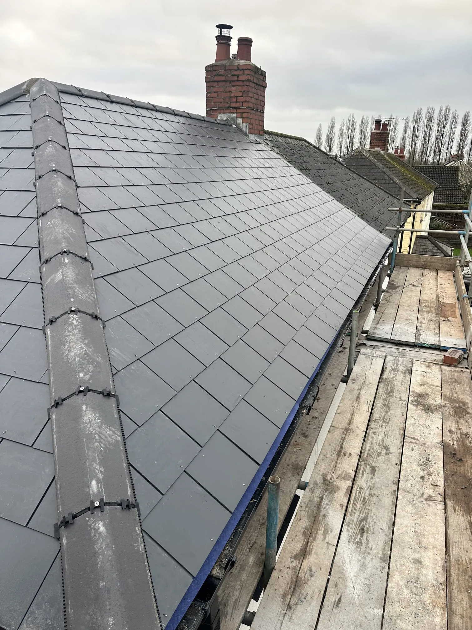 Close-up view of a recently installed residential roof featuring modern dark grey tiles and a new ridge system, with scaffolding for access.