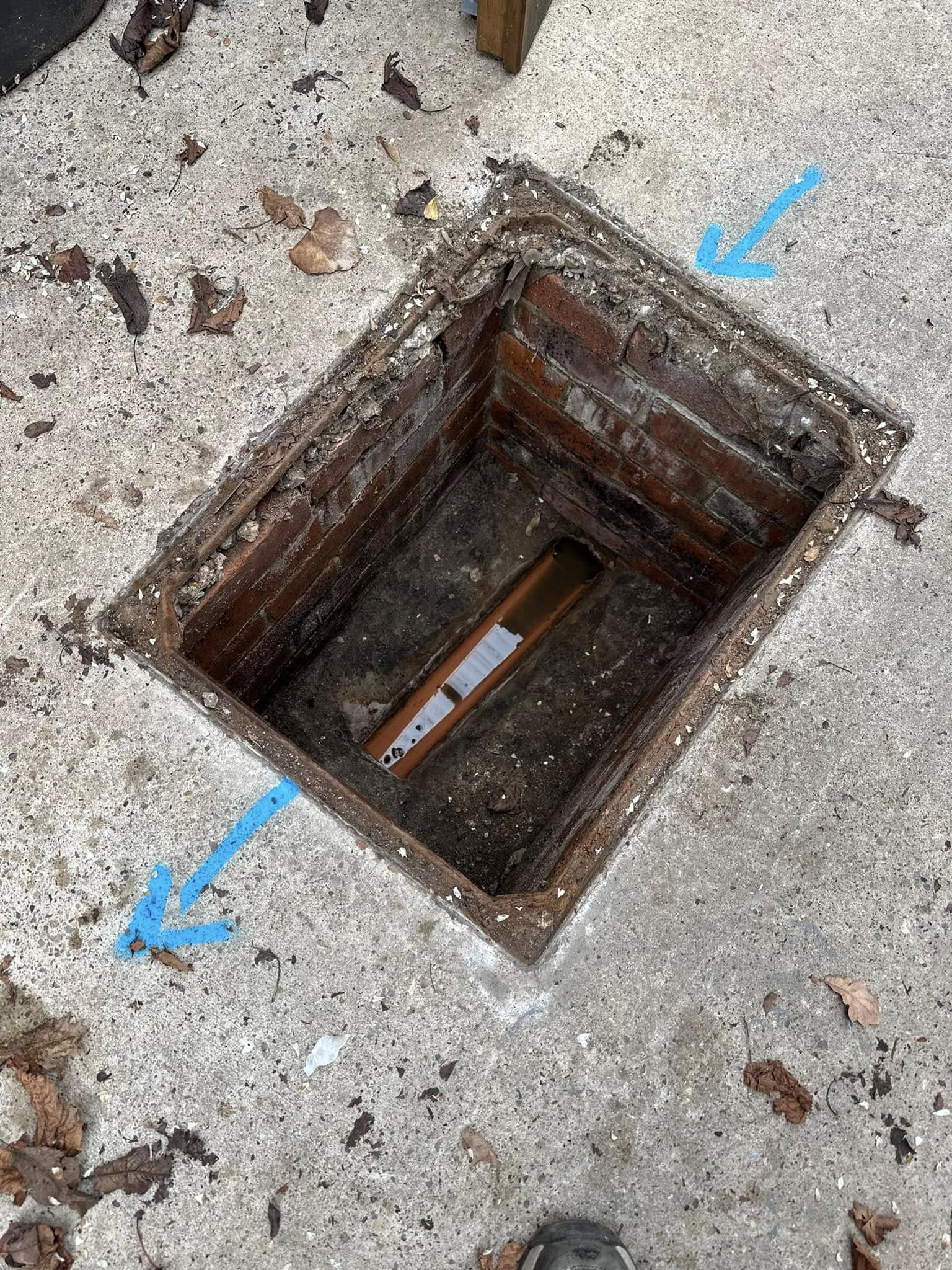 Open drainage inspection chamber or manhole with an orange pipe visible inside, marked with blue arrows on the concrete surround. Indicates a drain survey or repair in progress.