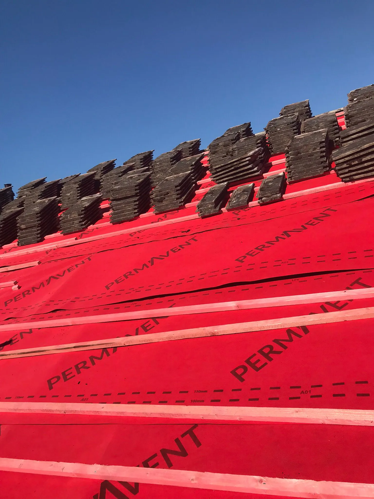 Roof installation in progress showing red underlayment, wooden battens, and stacked dark roof tiles under a clear blue sky.