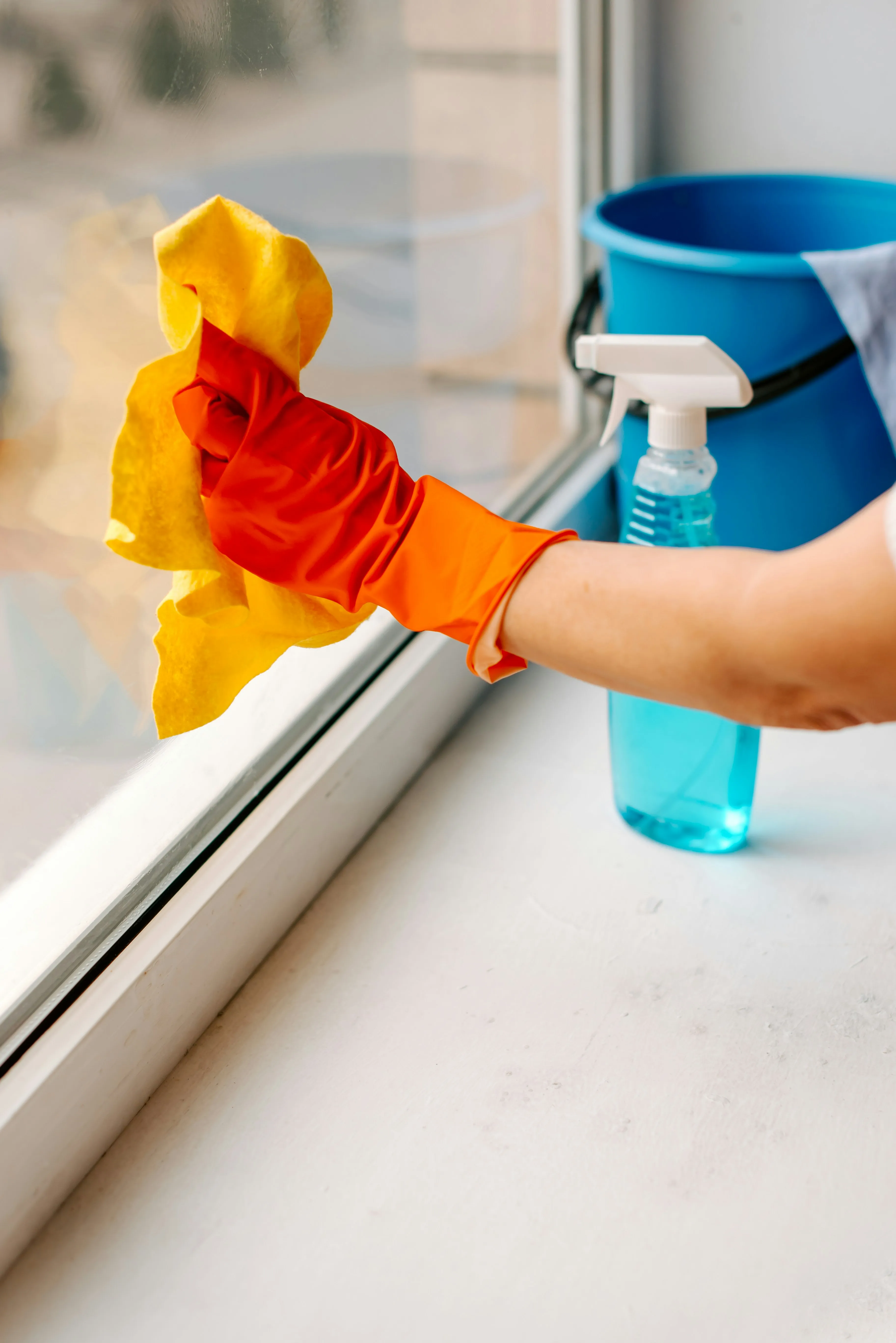 Person in orange glove cleaning a window with a yellow cloth and spray bottle