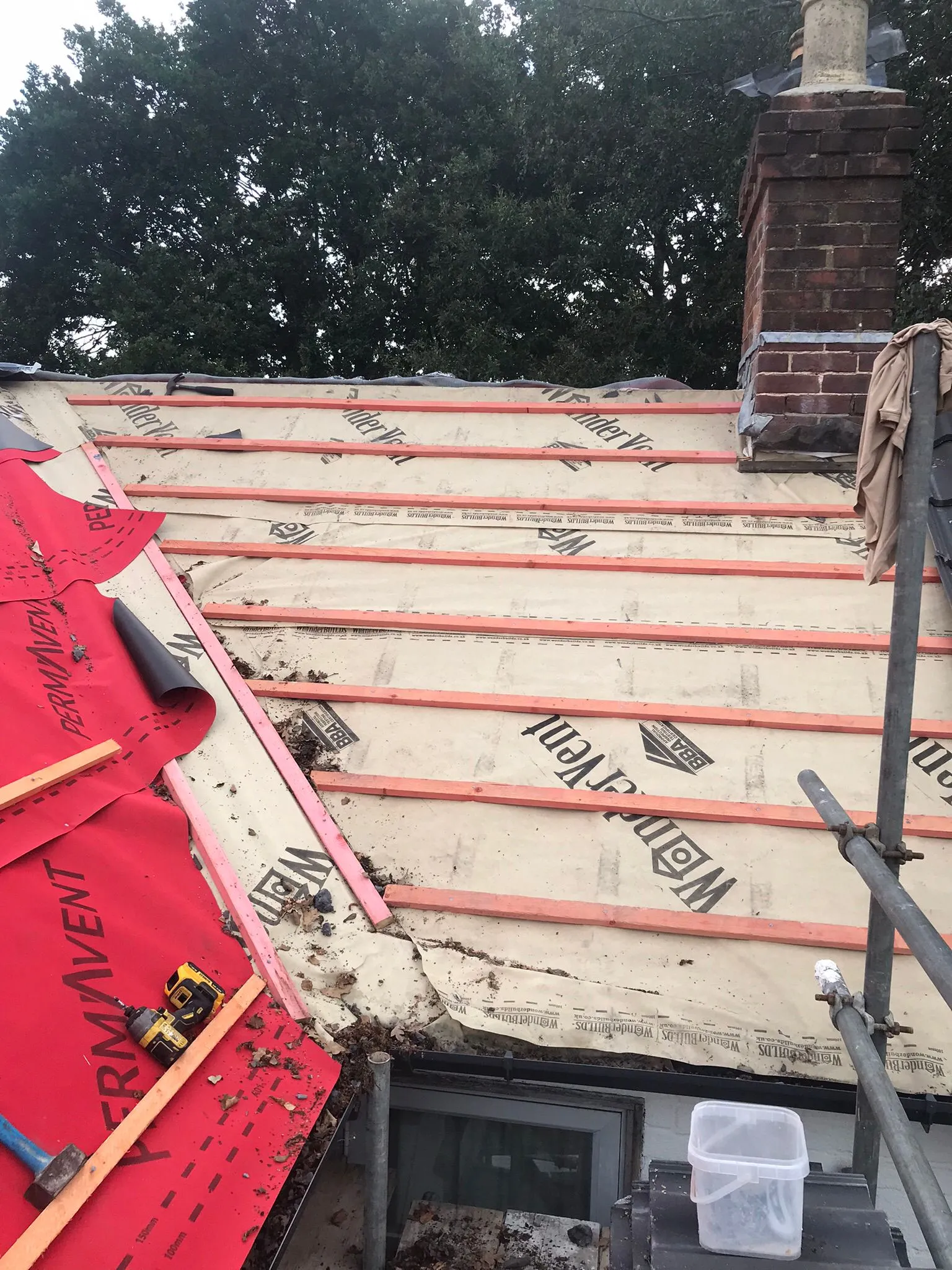 Pitched roof construction or re-roofing in progress, showing underlayment, timber battens, and a chimney stack.