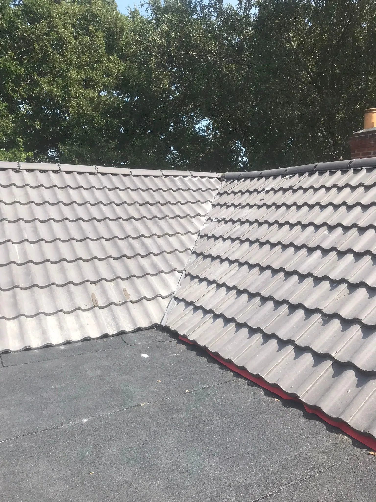 An image of a residential roof where a grey tiled pitched roof meets a dark flat roof, with trees and a chimney stack in the distance.