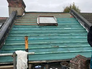 Pitched roof structure with green underlayment, wooden battens, a chimney, and a skylight window during a new roof installation.