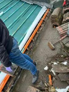 Roofer installing or replacing tiles on a pitched roof, with a green metal roof section and roofing debris.