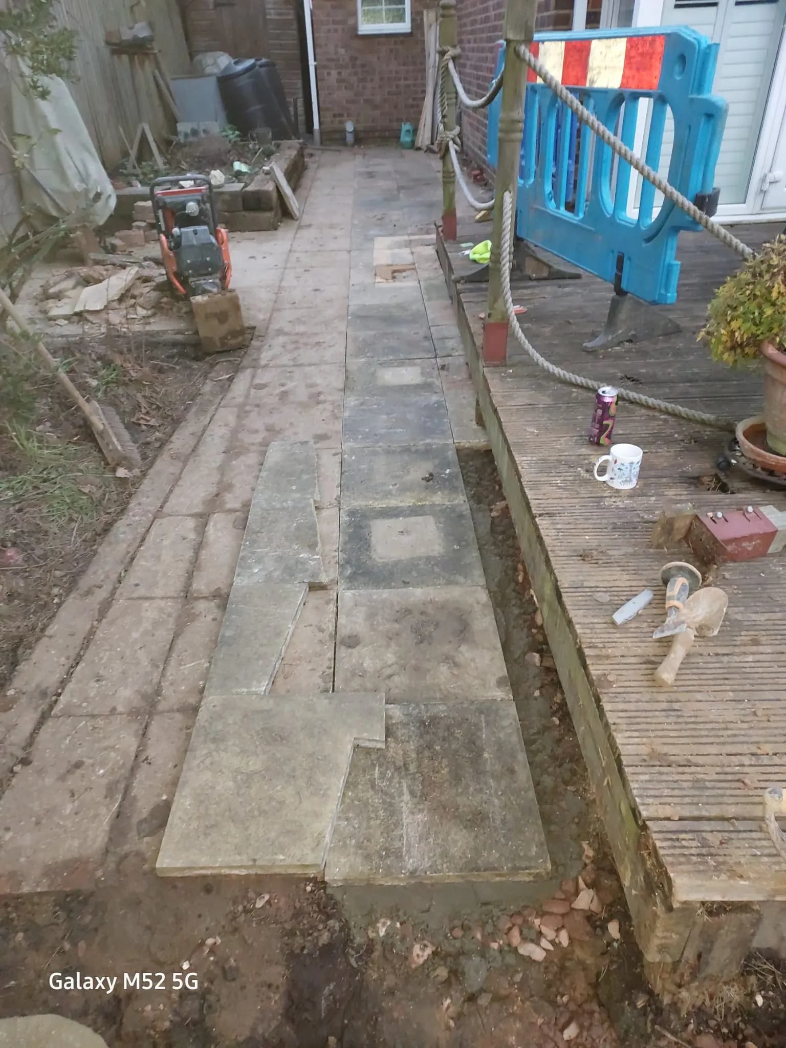 An outdoor area during drainage and paving work, featuring partially laid patio slabs, an open trench, and construction equipment like a plate compactor.