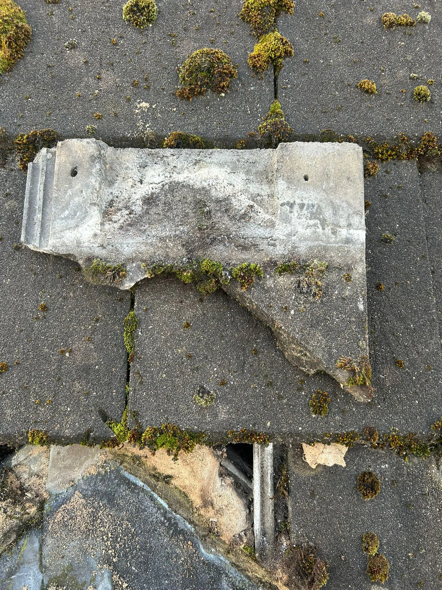 A close-up view of a dark grey roof with significant patches of green moss and a large, broken concrete roof tile exposing the roof's underlying structure.