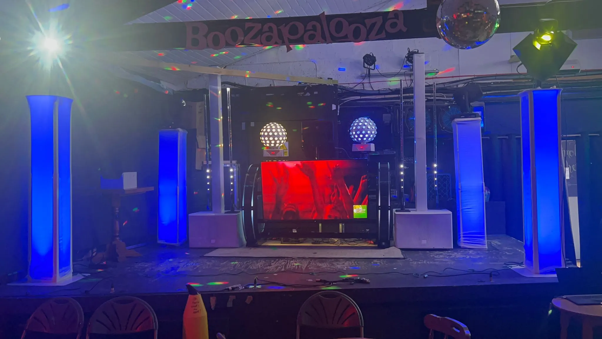 A professional DJ setup with a large screen, blue uplighting, disco balls, and speakers, ready for a party.