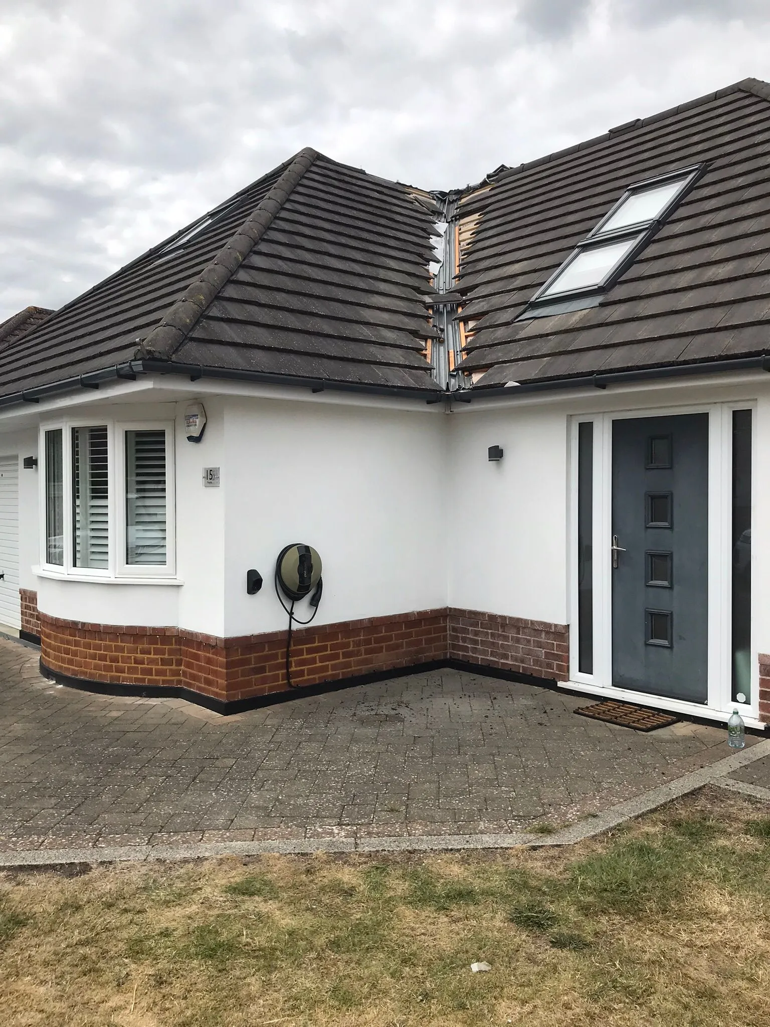 A modern house with a damaged or undergoing repair tiled roof, featuring exposed roof structure, two Velux skylights, and an electric vehicle charging station on the white rendered wall.