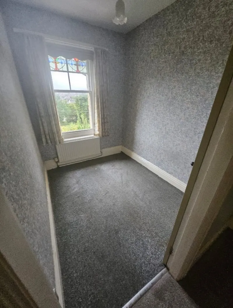 Empty room with grey carpet and wallpaper, radiator, and a window with stained glass details, showcasing a property cleared by Llandudno Rhos on sea House Clearance & Rubbish Removals.