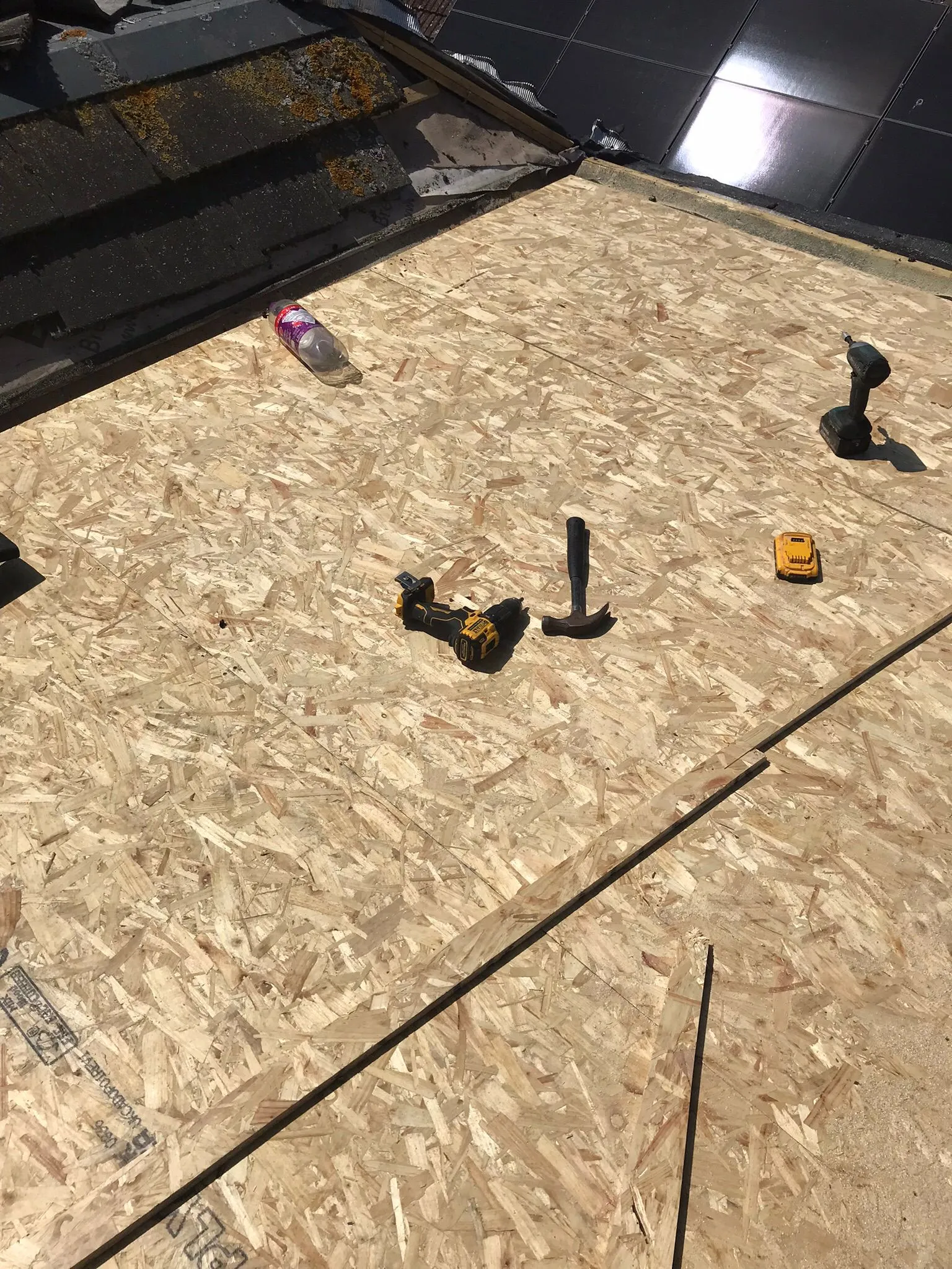 OSB sheathing laid on a roof under construction, with various roofing tools scattered on the surface and existing roof sections with solar panels and moss-covered tiles in the background.