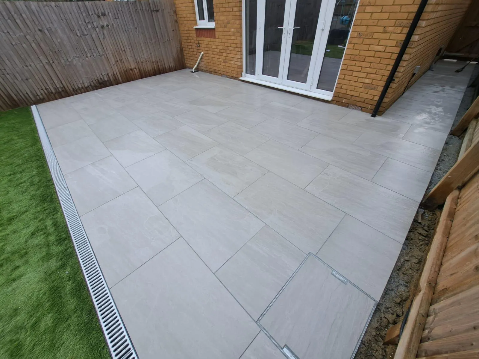 A newly installed light grey patio with modern tiles, a linear drainage channel, and artificial grass next to a house.