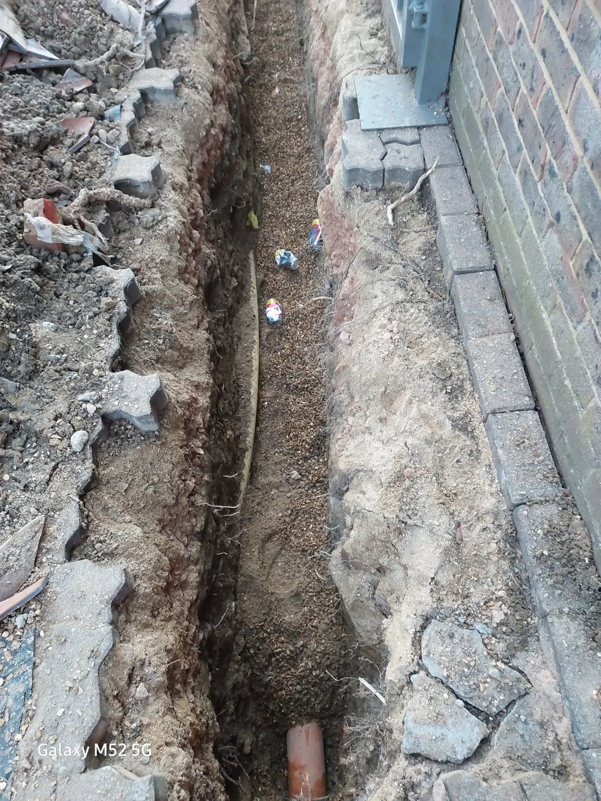 A construction trench with exposed drainage pipe and gravel, indicating ongoing drain repair or new installation.