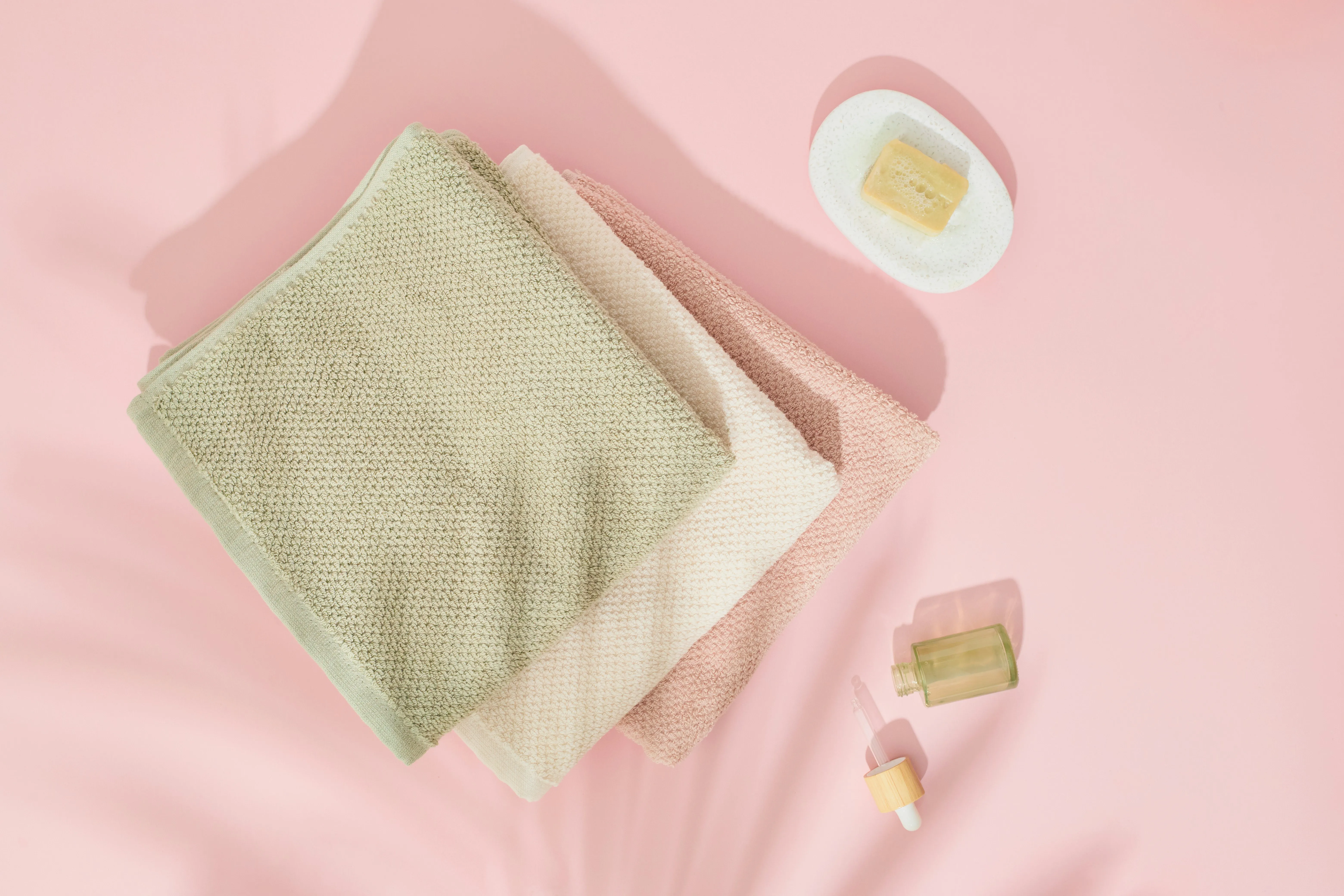 Eco-friendly cleaning supplies with folded soft towels, natural bar soap, and a dropper bottle on a pink background.