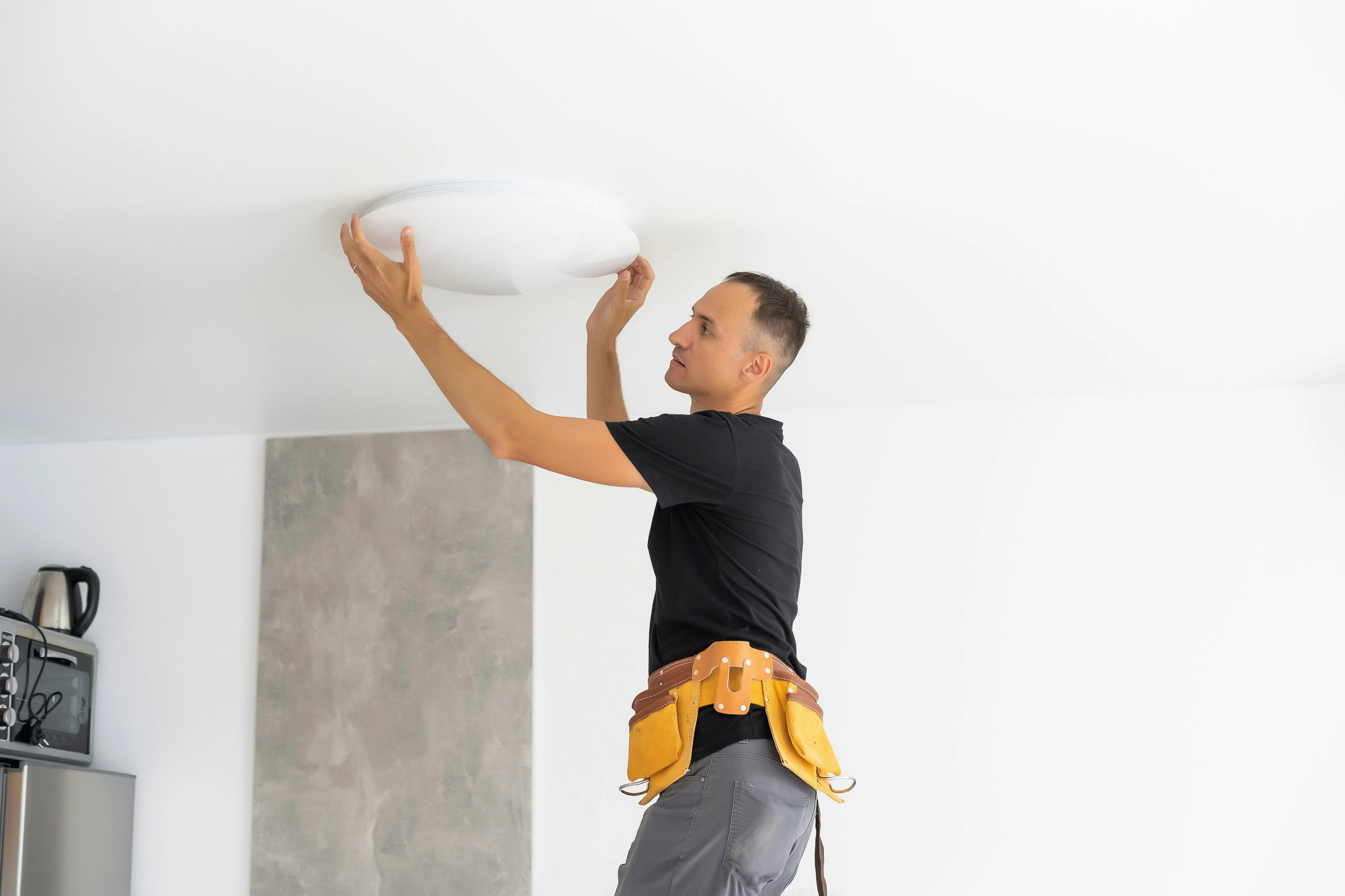 An electrician wearing a tool belt, installing or repairing a circular LED light fixture on a white ceiling.